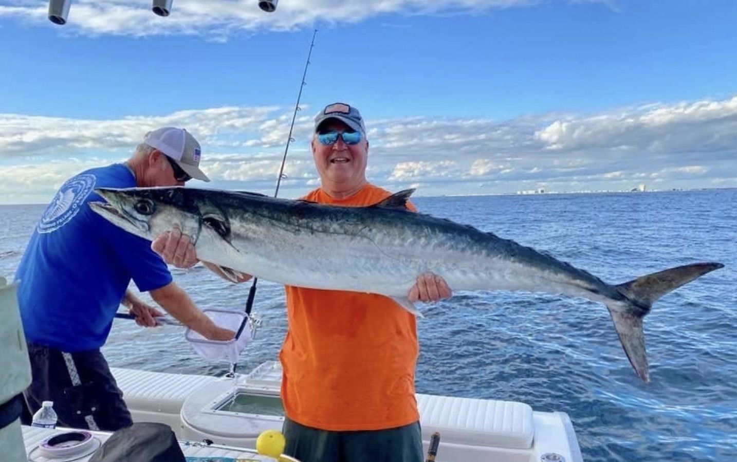 Two people on a boat hold a large fish over the ocean under a bright blue sky.
