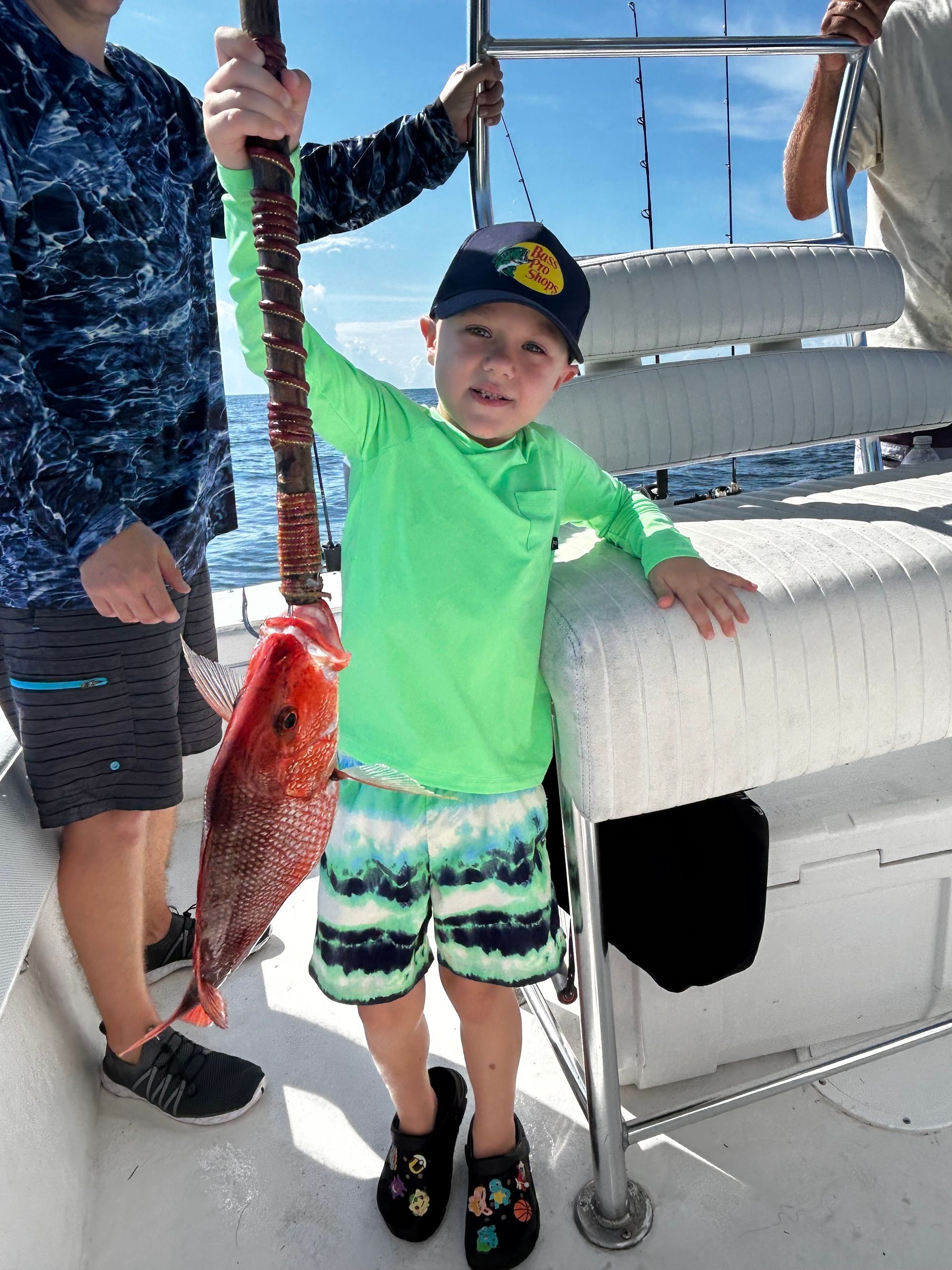 Child holding up a red fish on a boat, wearing a bright green shirt and black cap.