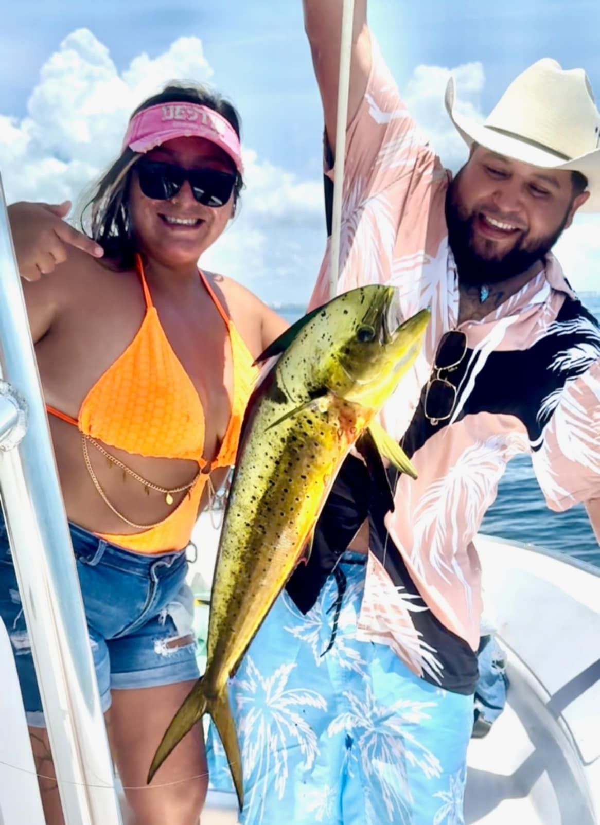 Two people on a boat holding a large colorful fish, smiling outdoors under a blue sky.