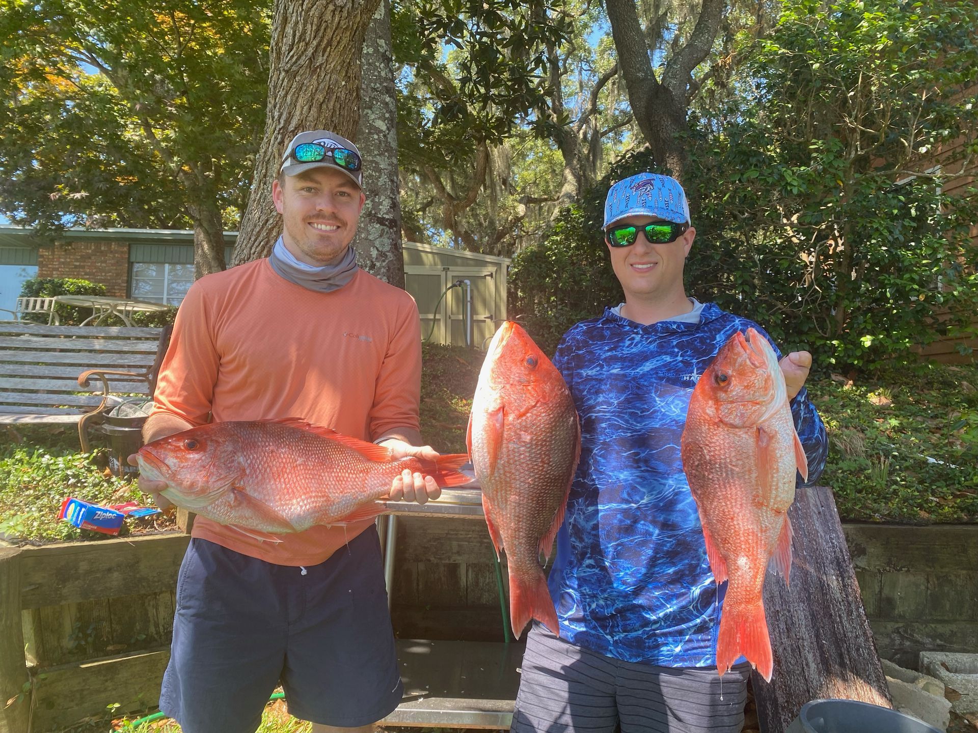 Two people holding large fish outdoors by a lakeside, standing in front of trees and a dock.