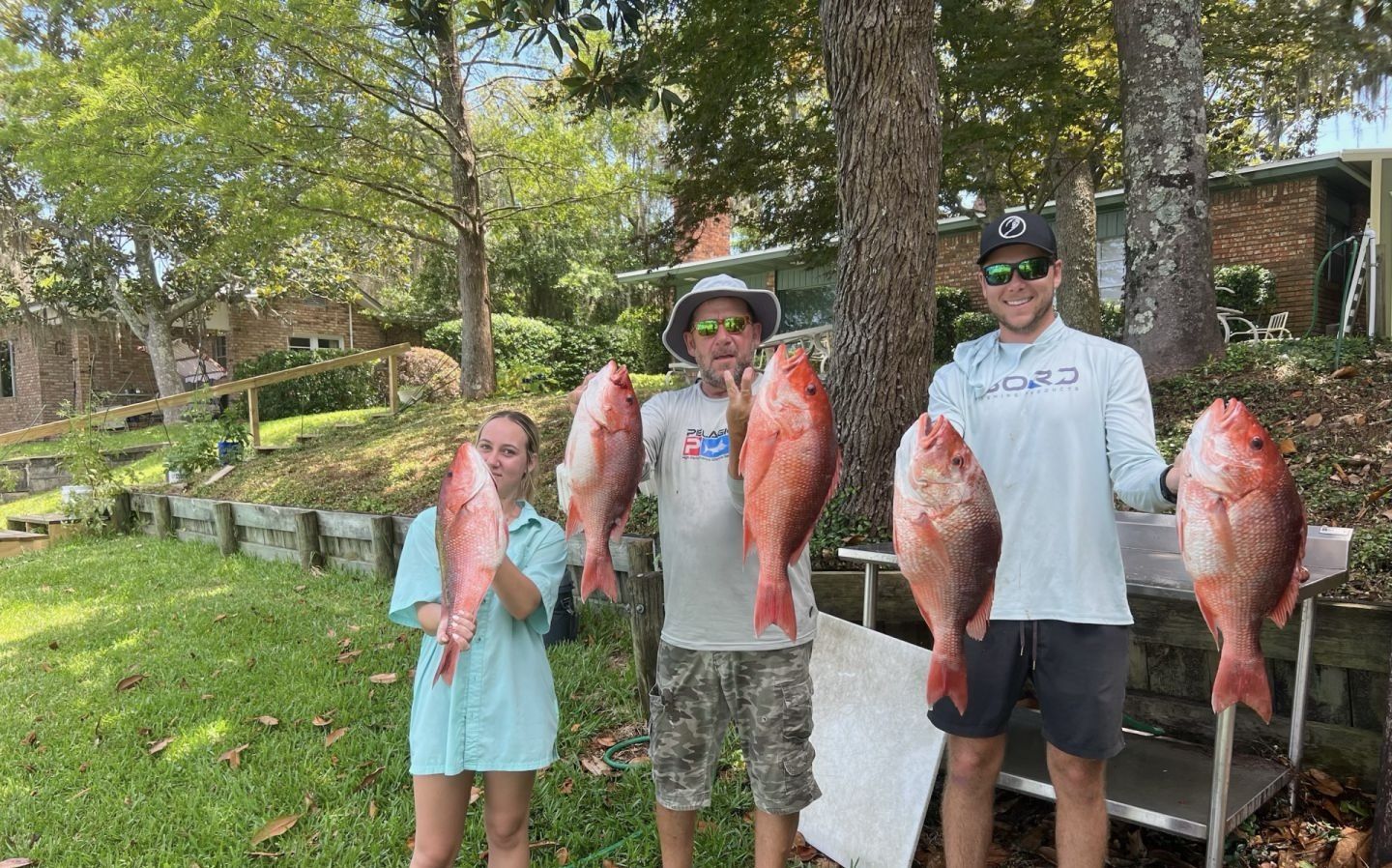 Three anglers holding large red fish outdoors by a wooded riverbank