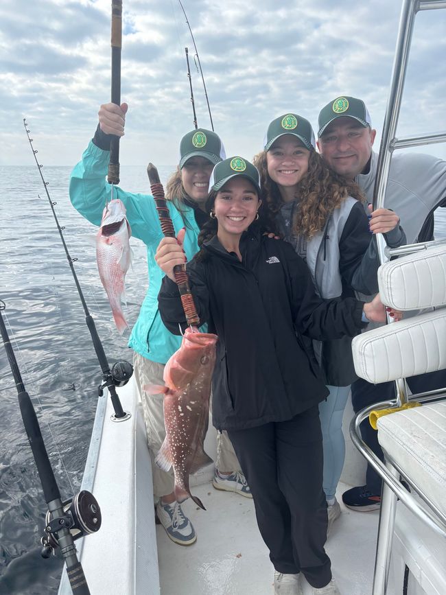Four people smiling on a boat, holding fishing rods and caught fish on a cloudy day.