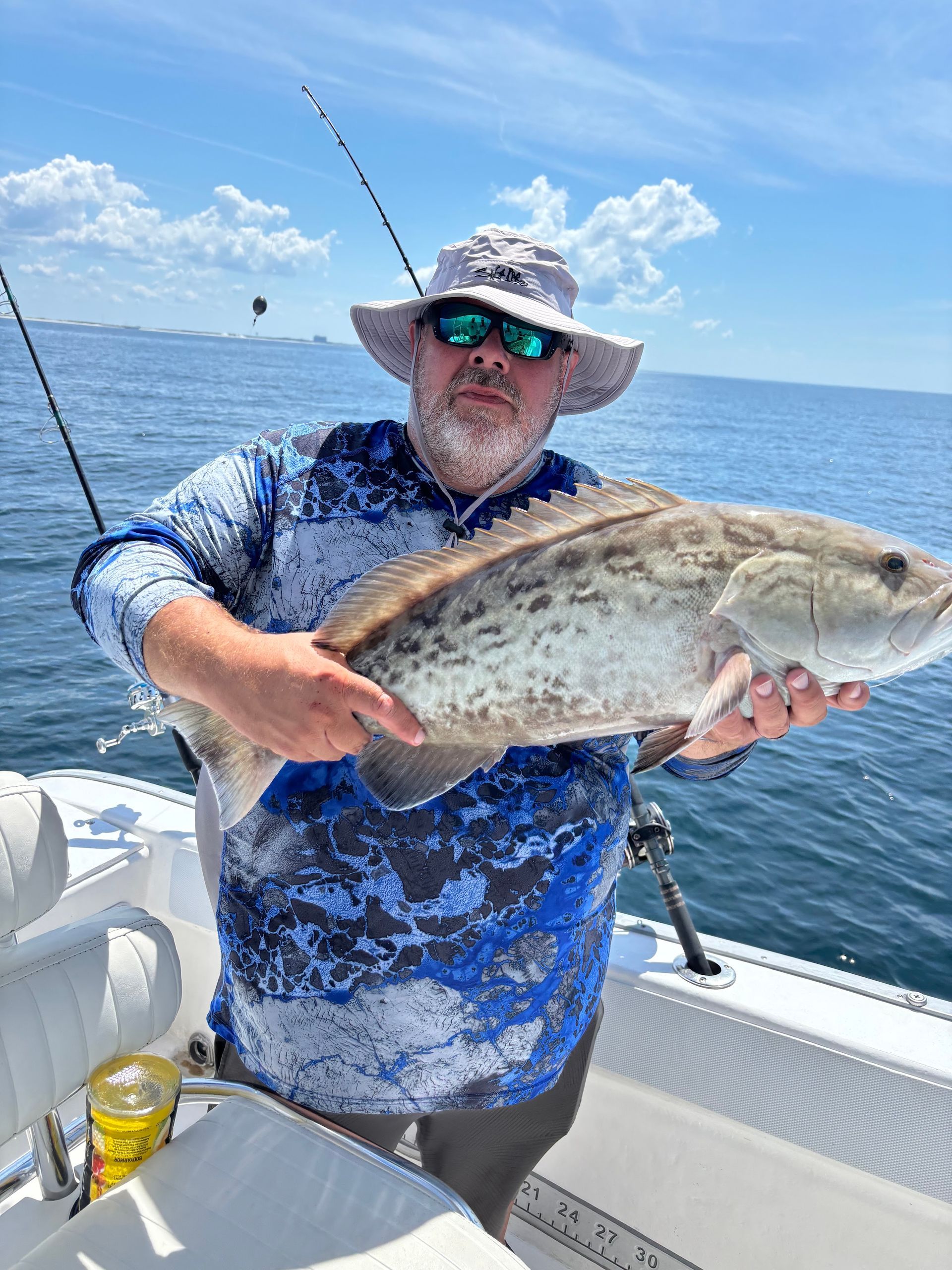 Man holding a large fish on a boat at sea, wearing a blue shirt, hat, and sunglasses.