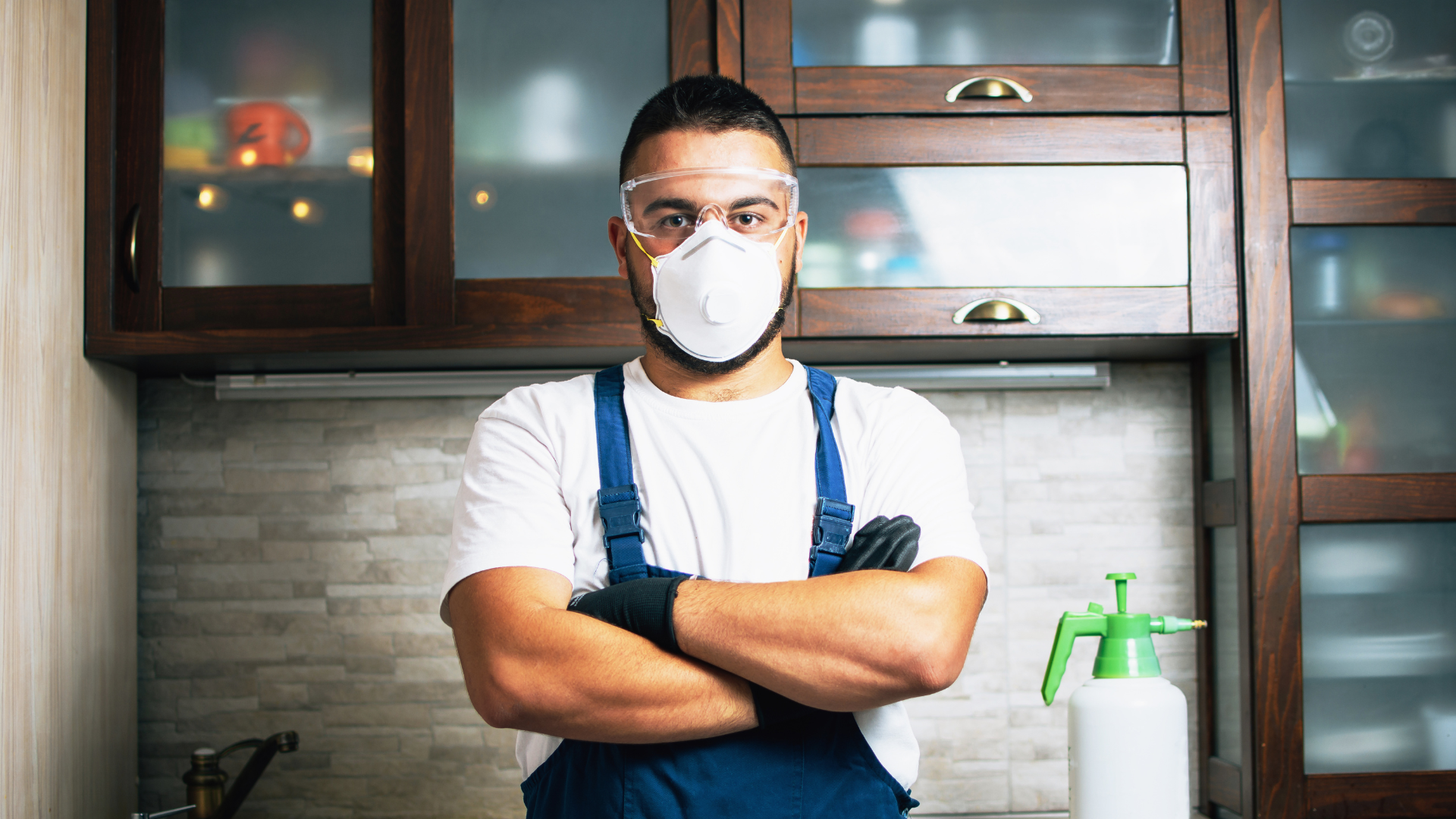 A man wearing a mask and goggles is standing in a kitchen with his arms crossed.