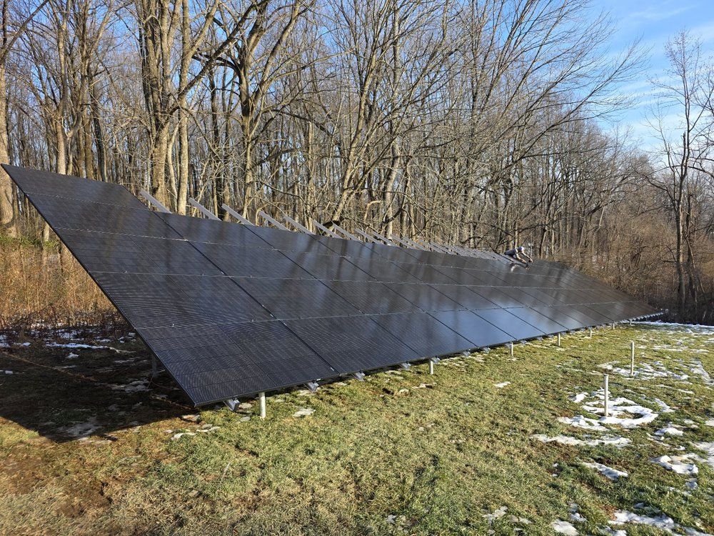 Large tilted solar panel array in a wooded field with patches of snow under a clear sky