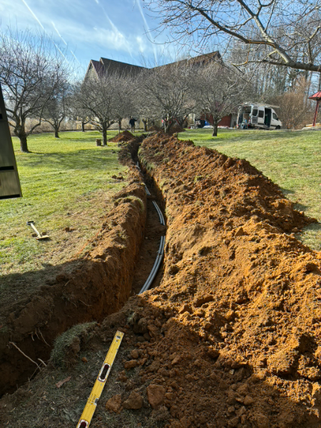 Deep trench dug through a grassy yard, with piled dirt and a house in the background.