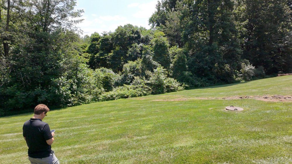 Person standing in a grassy field, looking toward trees and a wooded hillside.