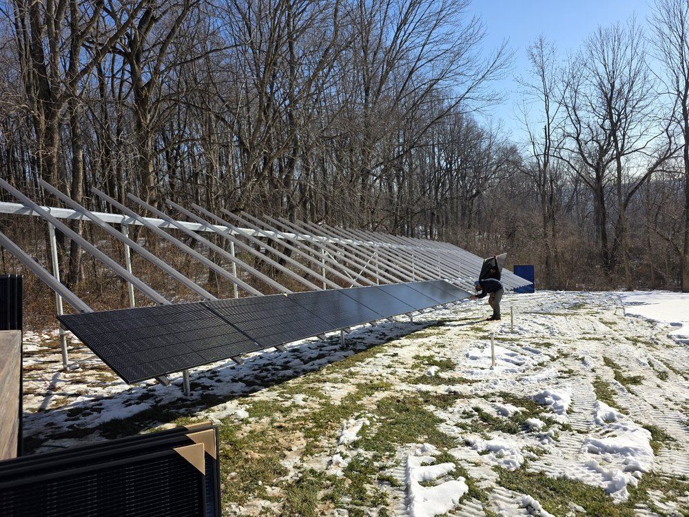 Large solar panel array in a snowy field with a person walking beside it under bare trees.