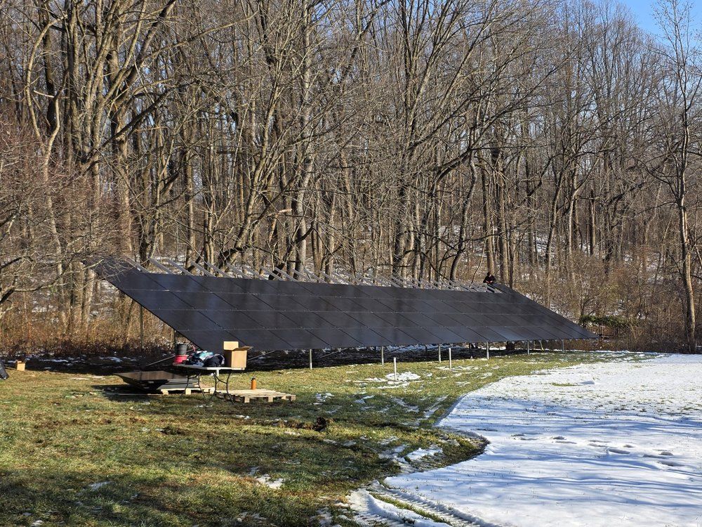 Solar panel array in a grassy field beside a snowy edge, with bare trees in the background.