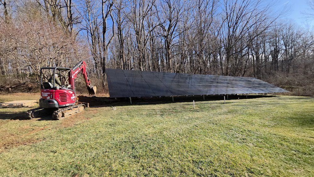 Red excavator lifting a long black metal panel in a grassy field with bare trees in the background