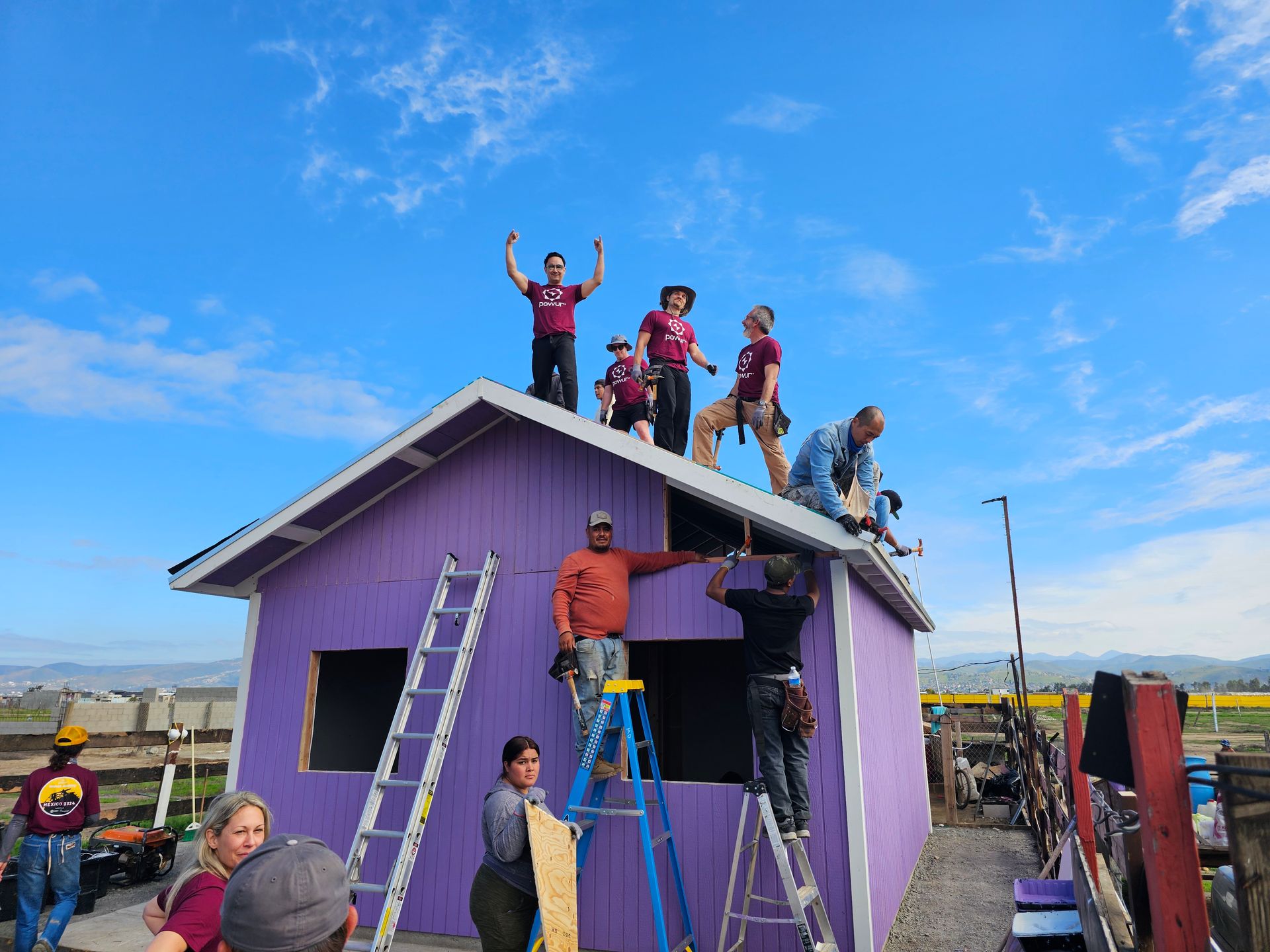Workers on a purple house roof and ground, painting and repairing under a bright blue sky.