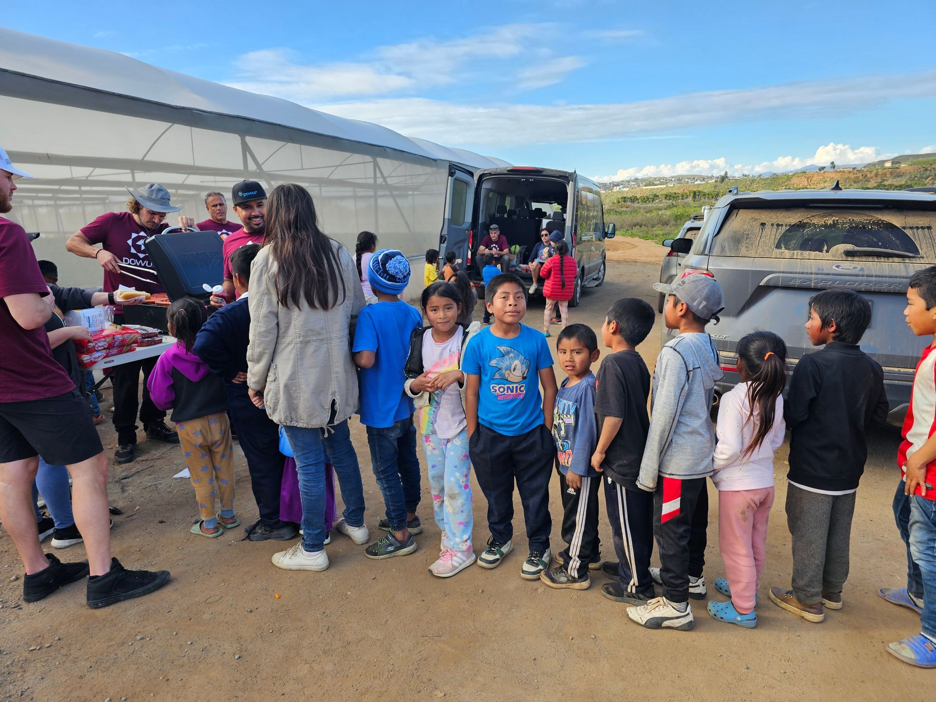 Children and adults gathered outdoors near a trailer and SUV, waiting in line beside a table under a clear sky.