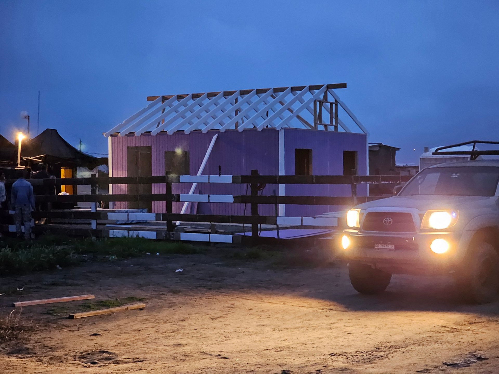 Partially built house at dusk with a pickup truck headlights on in front of a wooden fence