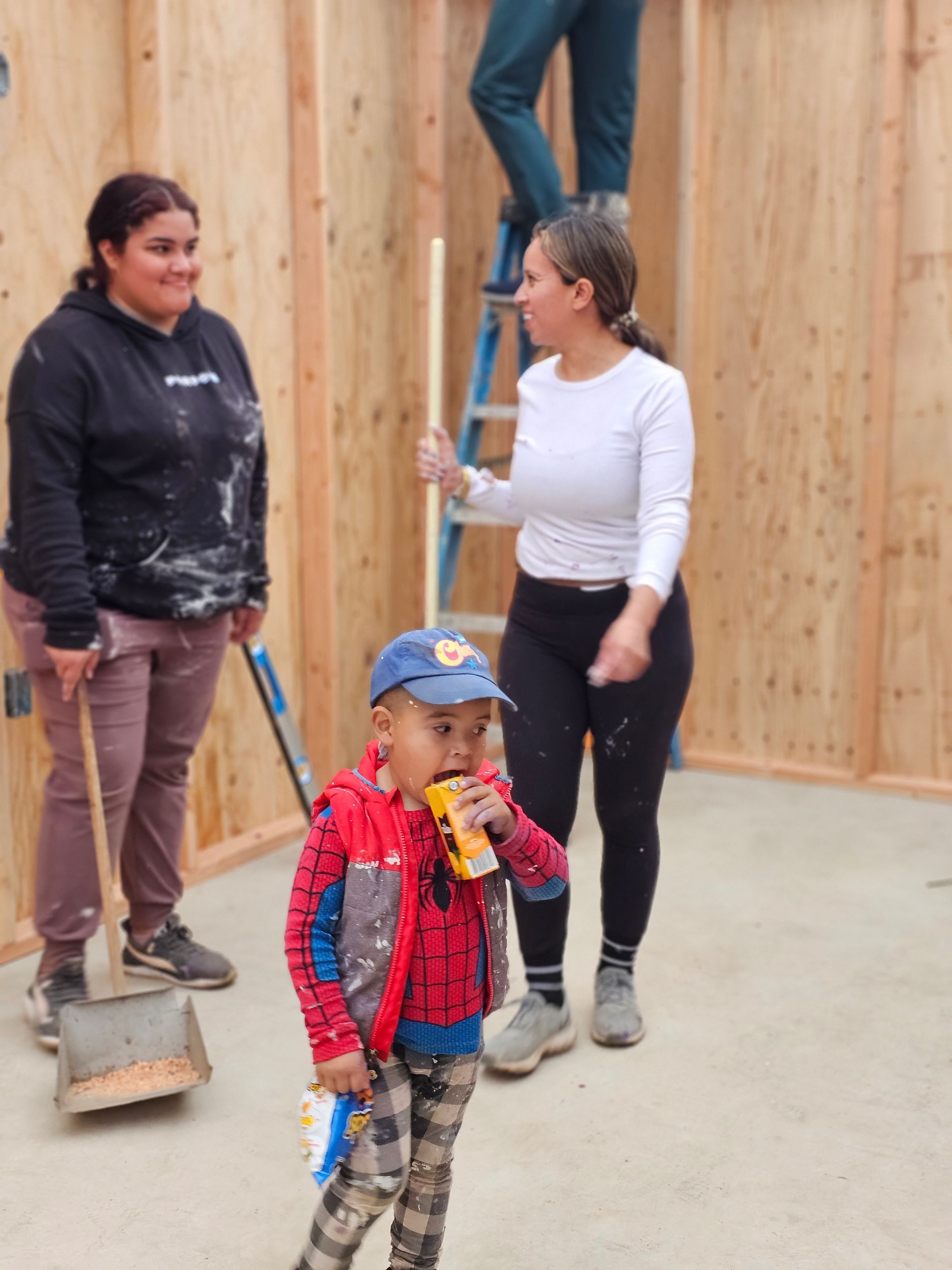 Three people standing in a wooden-walled room, one child in front drinking from a can.