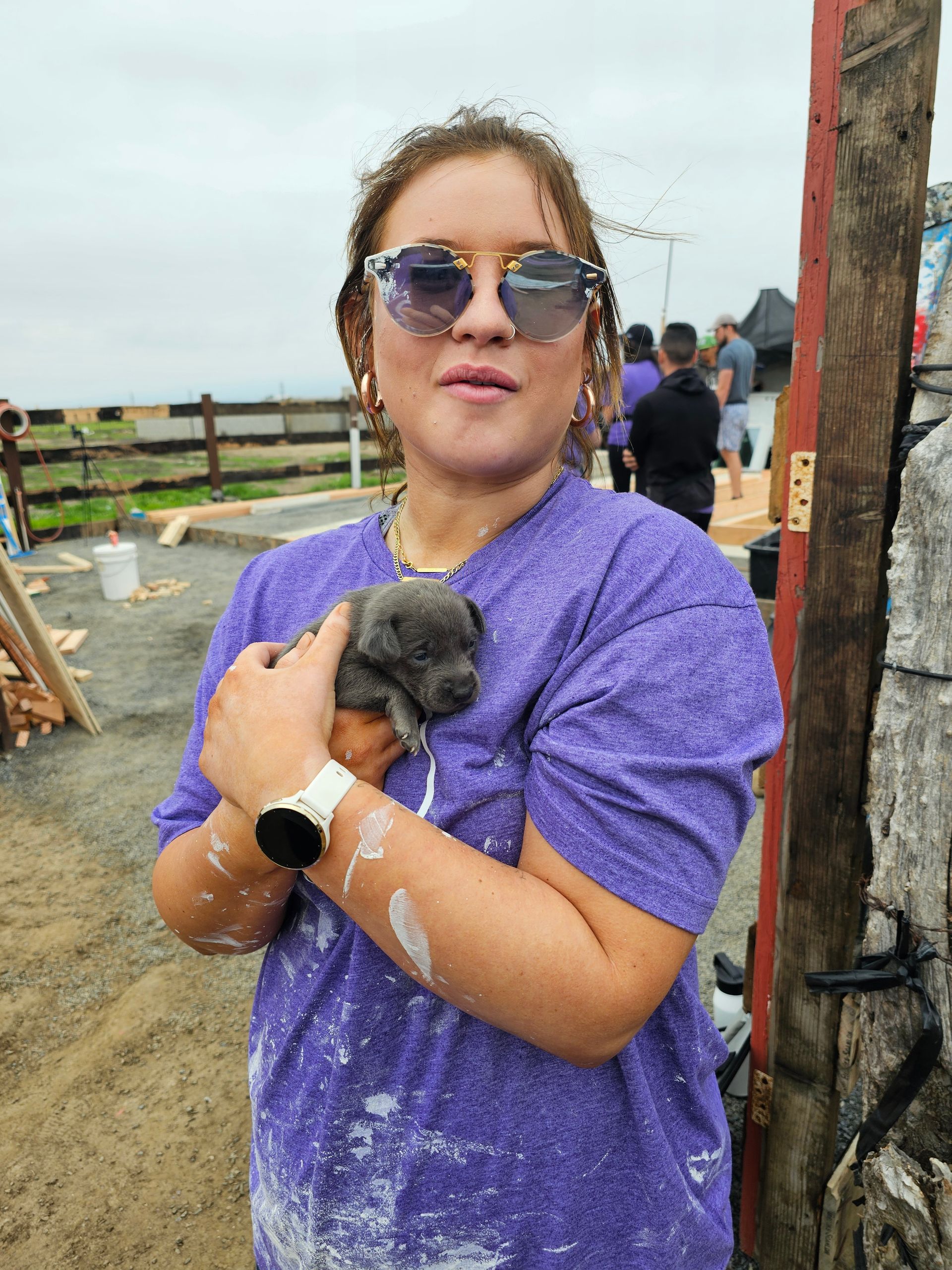 Woman in a purple shirt holding a small puppy outdoors near a fence on a muddy path.