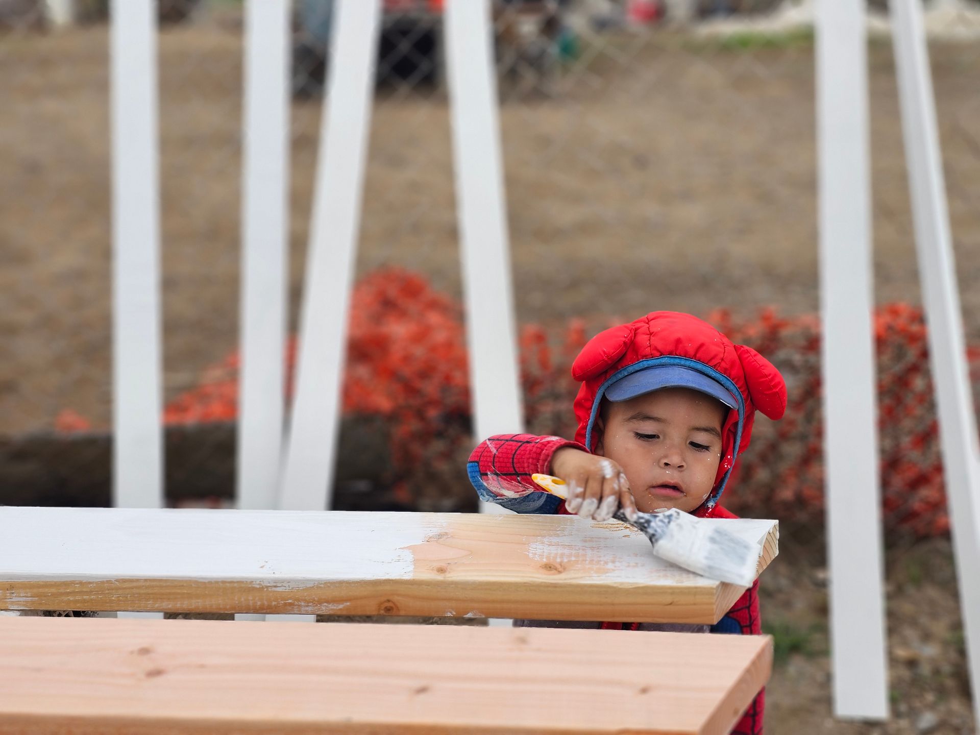 Child in a red winter hat leaning over a white bench outdoors, focused on a paper-covered surface