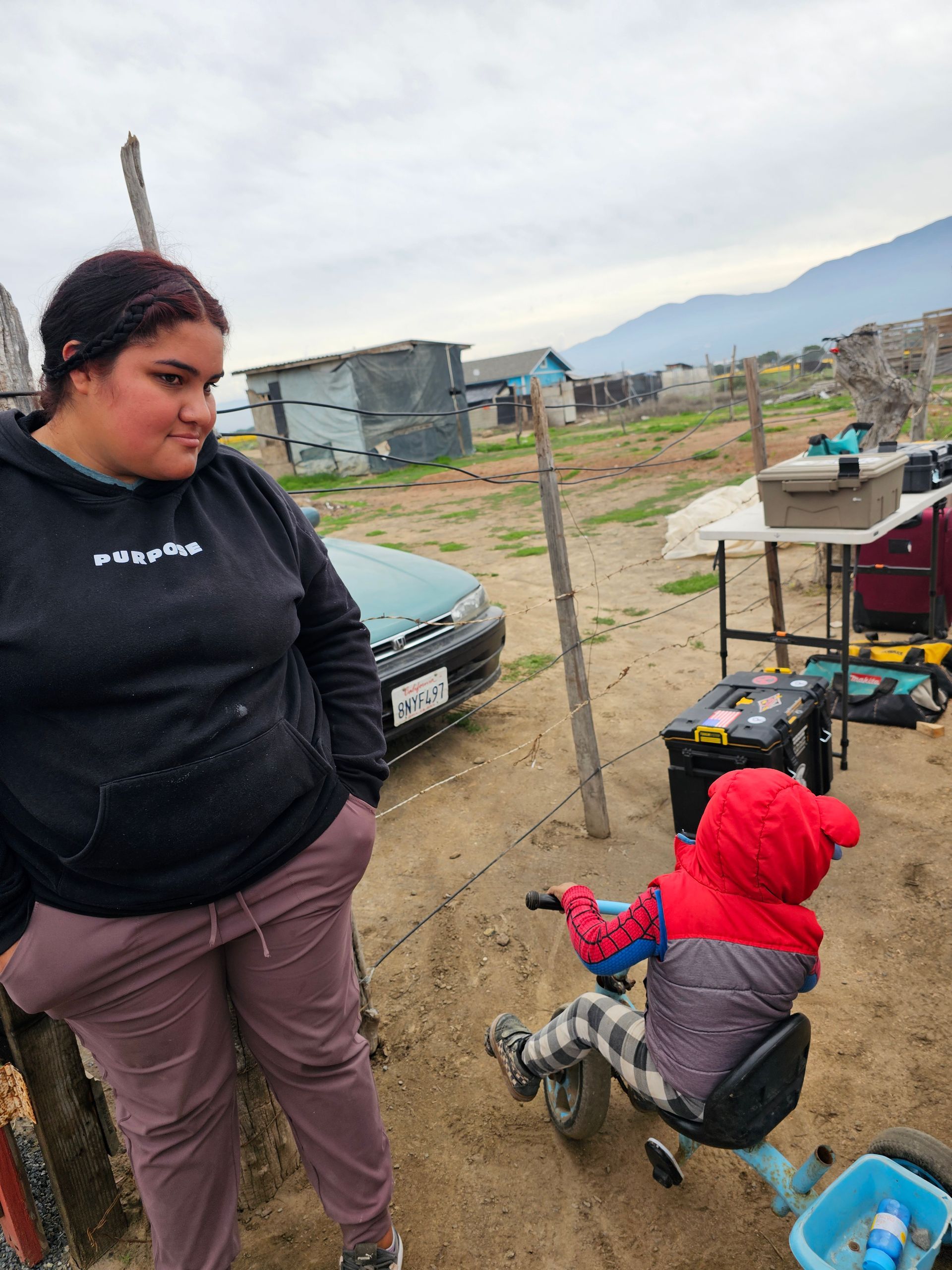 Person in black sweatshirt standing near child in red hoodie playing by a cart outdoors on a cloudy day