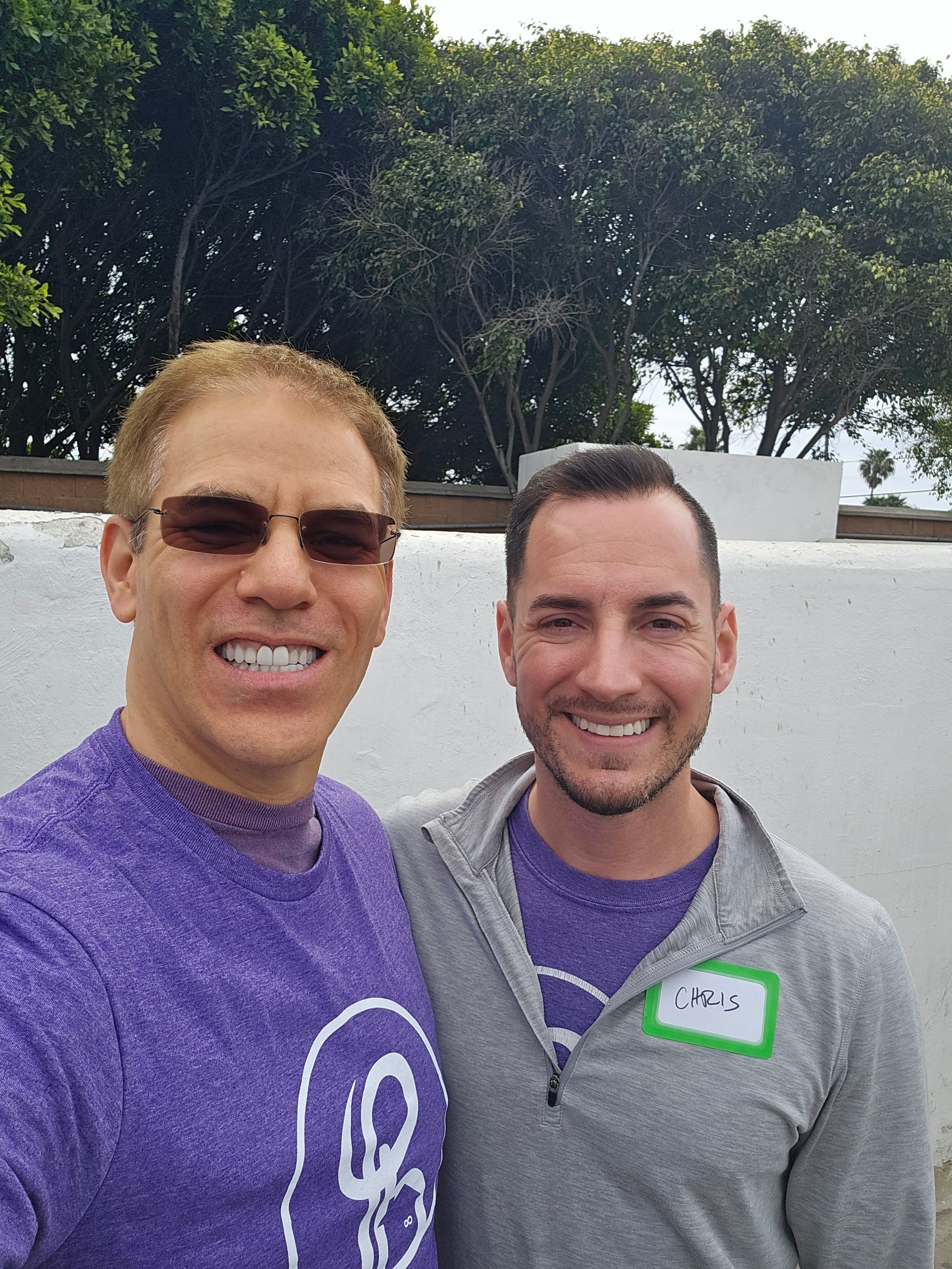 Two smiling men posing outdoors, one in a purple shirt and sunglasses, the other in a gray hoodie.