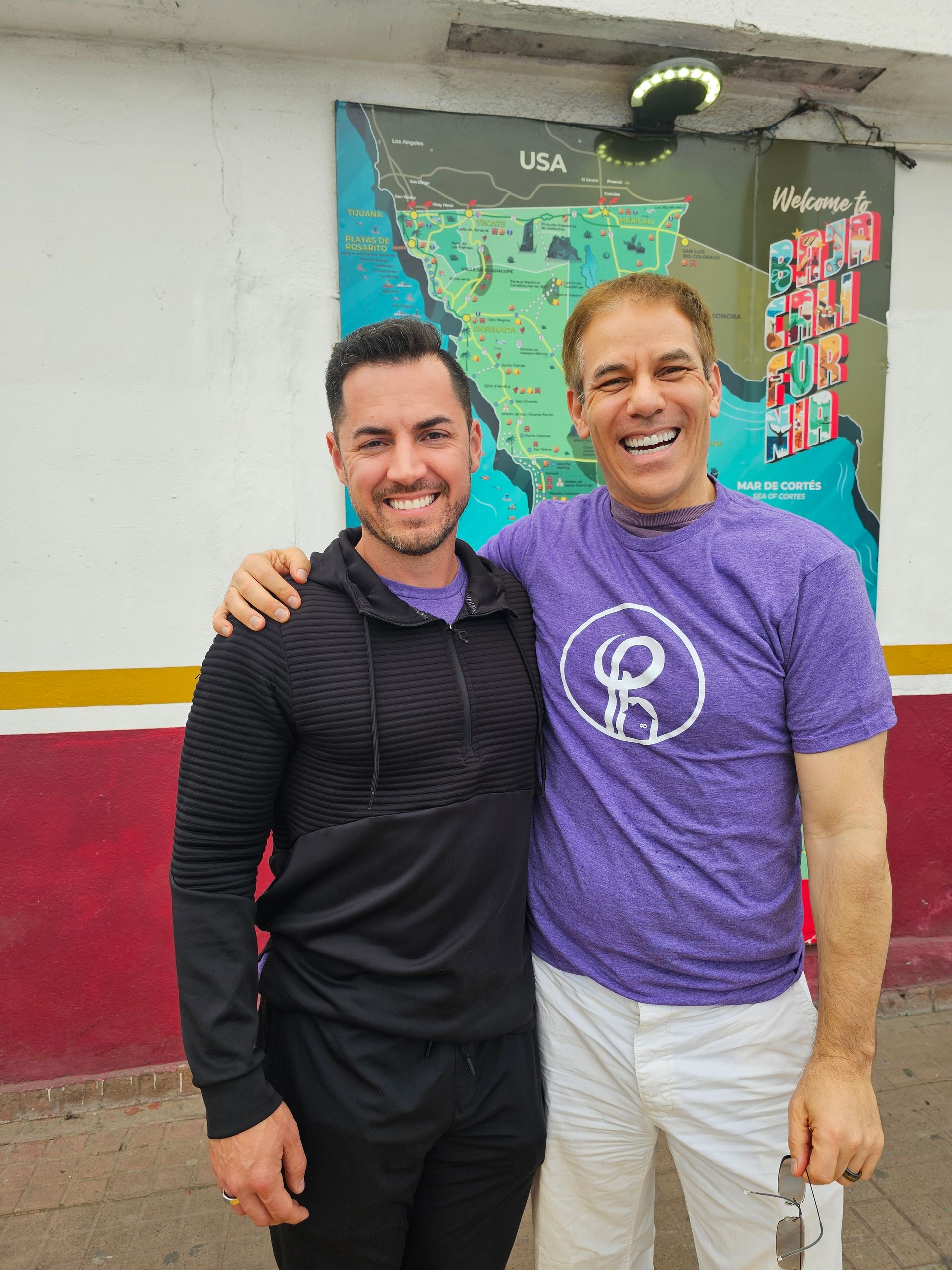Two smiling men stand together in front of a colorful wall mural.