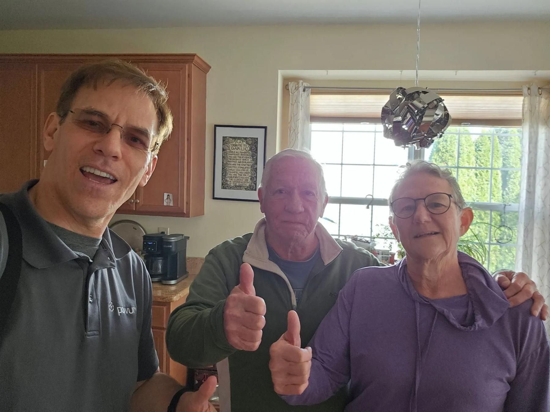 Three people smiling, giving thumbs up indoors near a window. 