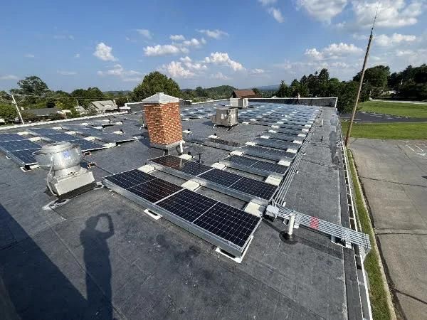 Rooftop solar panel array on a commercial building under a blue sky, with a brick chimney and other equipment.