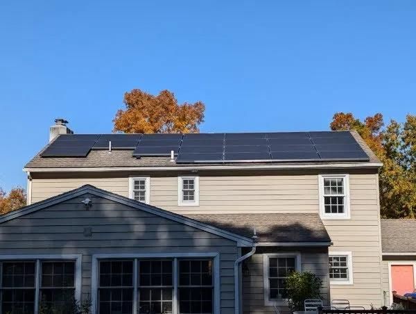 Solar panels installed on a residential roof. Panels are black, sky is blue, autumn trees in background.