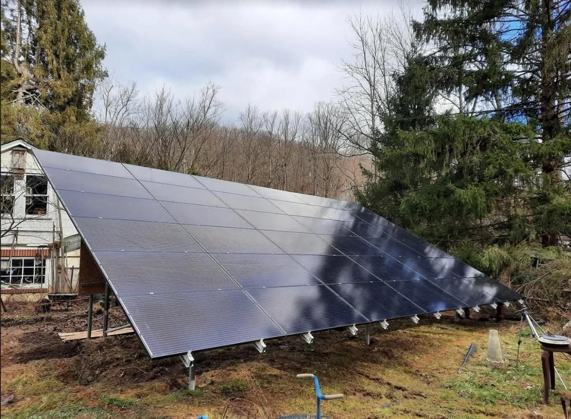 Solar panel array on a grassy hillside, tilted towards the viewer, against a backdrop of trees and a cloudy sky.