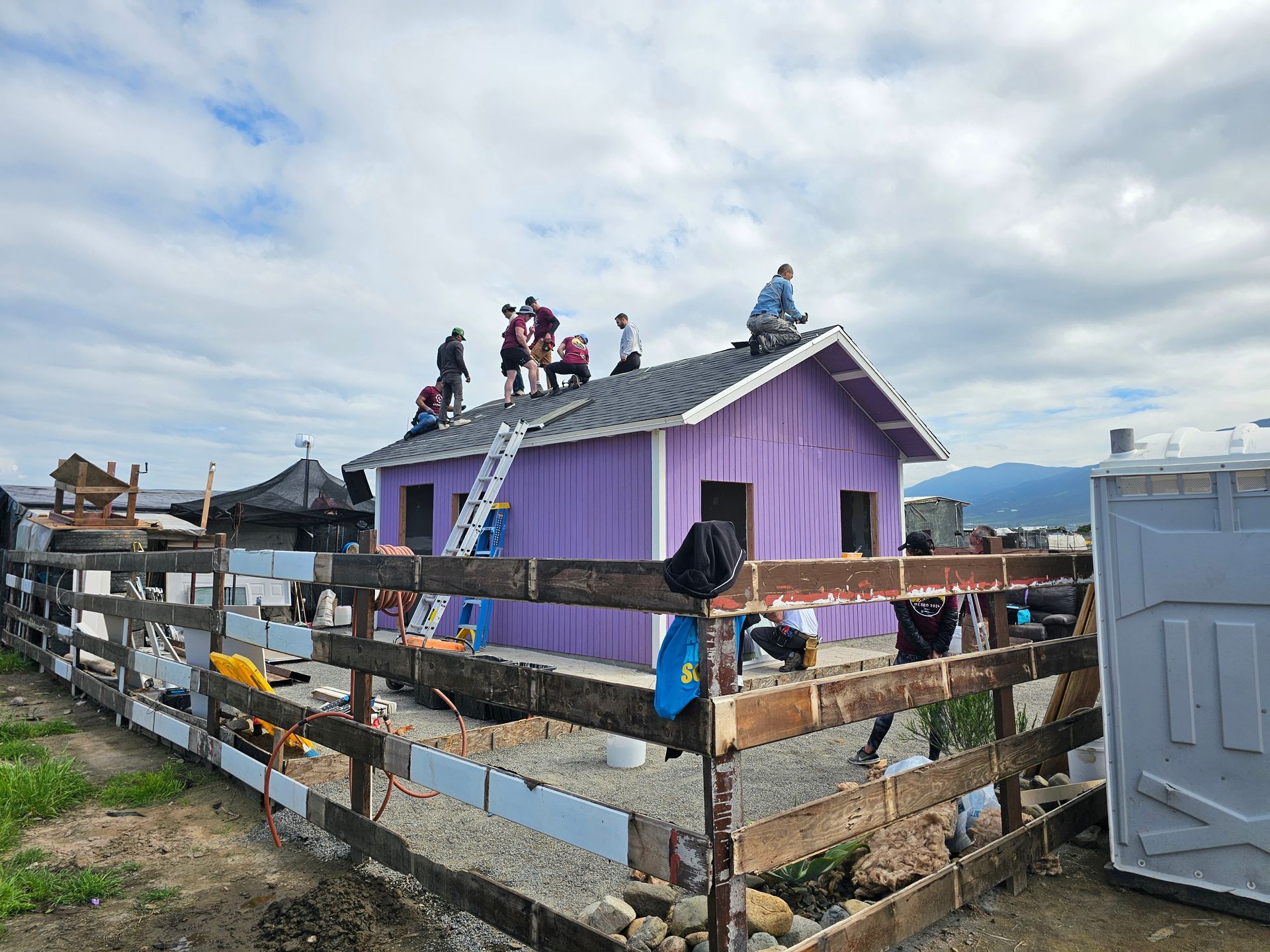 People on the roof of a purple house by a fenced waterfront, with a cloudy sky overhead.
