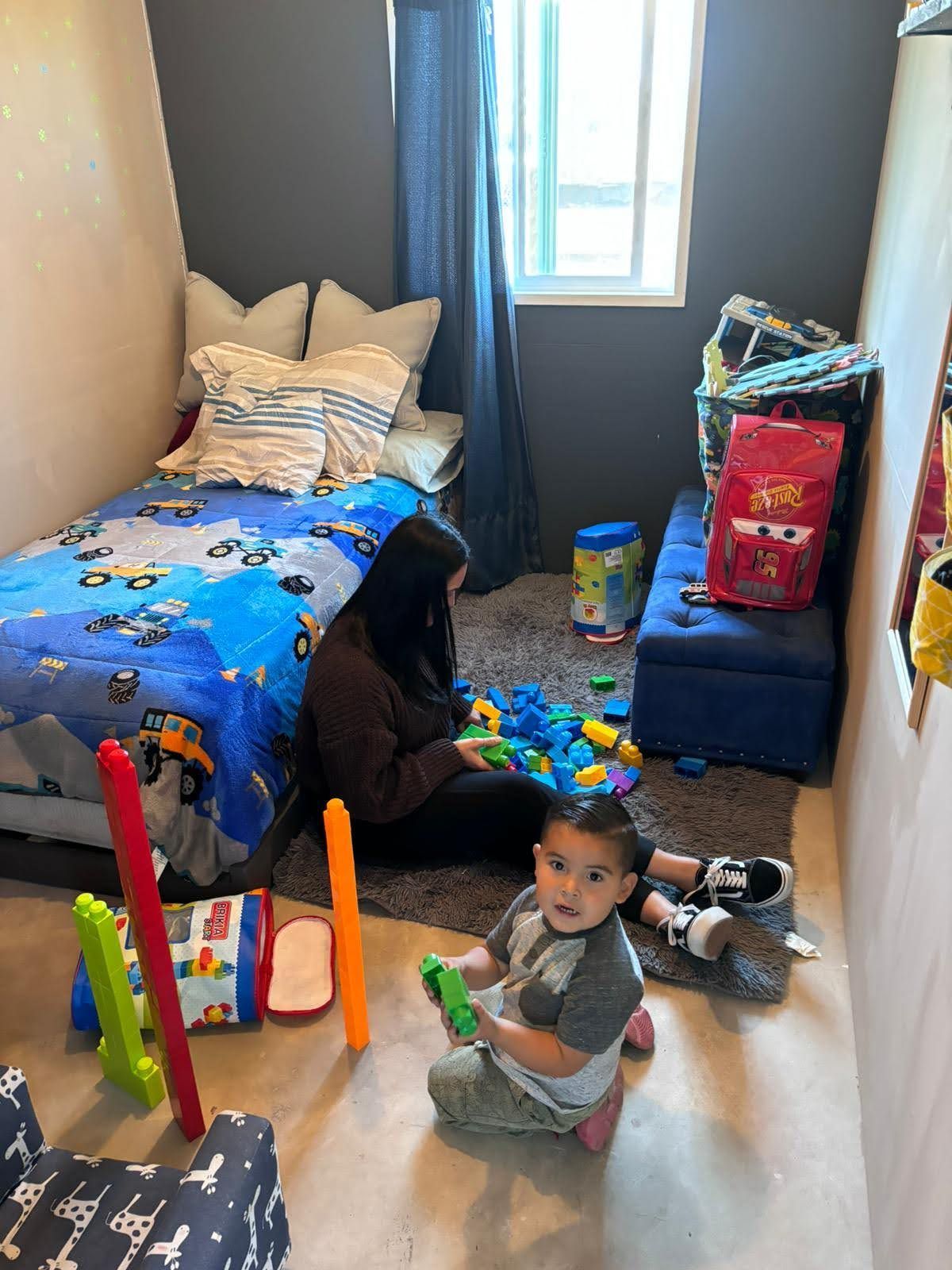 Child sitting on a bedroom floor with toys beside a bed, window, and stuffed animals.