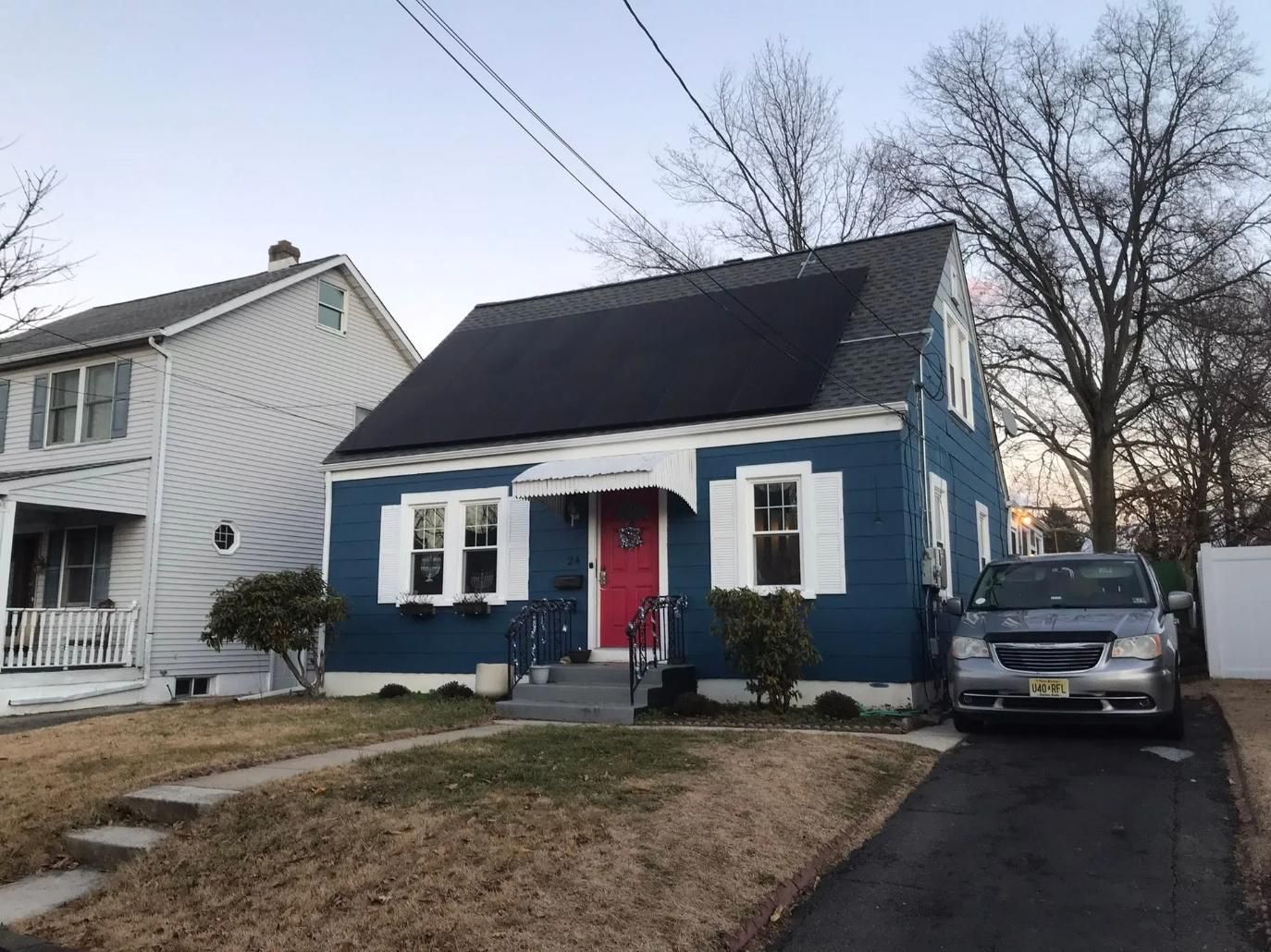 Blue house with black solar panels, red door, white shutters, parked silver car.