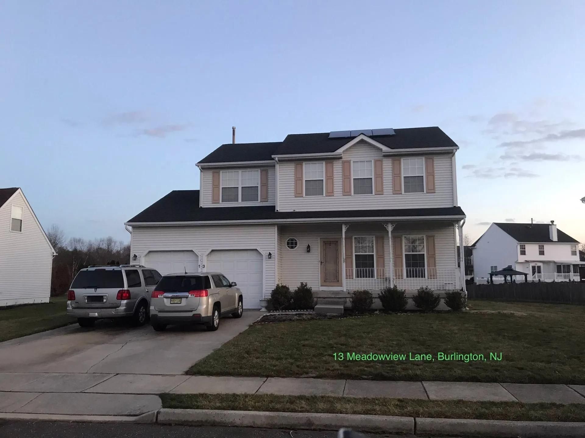 Two-story house with a black roof, beige siding, and three cars parked in the driveway; cloudy sky.