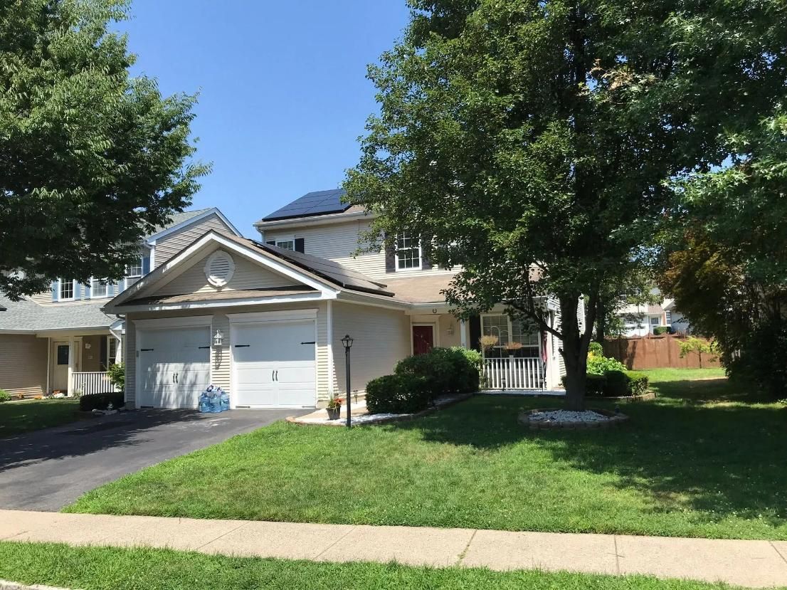 Two-story house with solar panels on roof, two-car garage, green lawn, and trees on a sunny day.