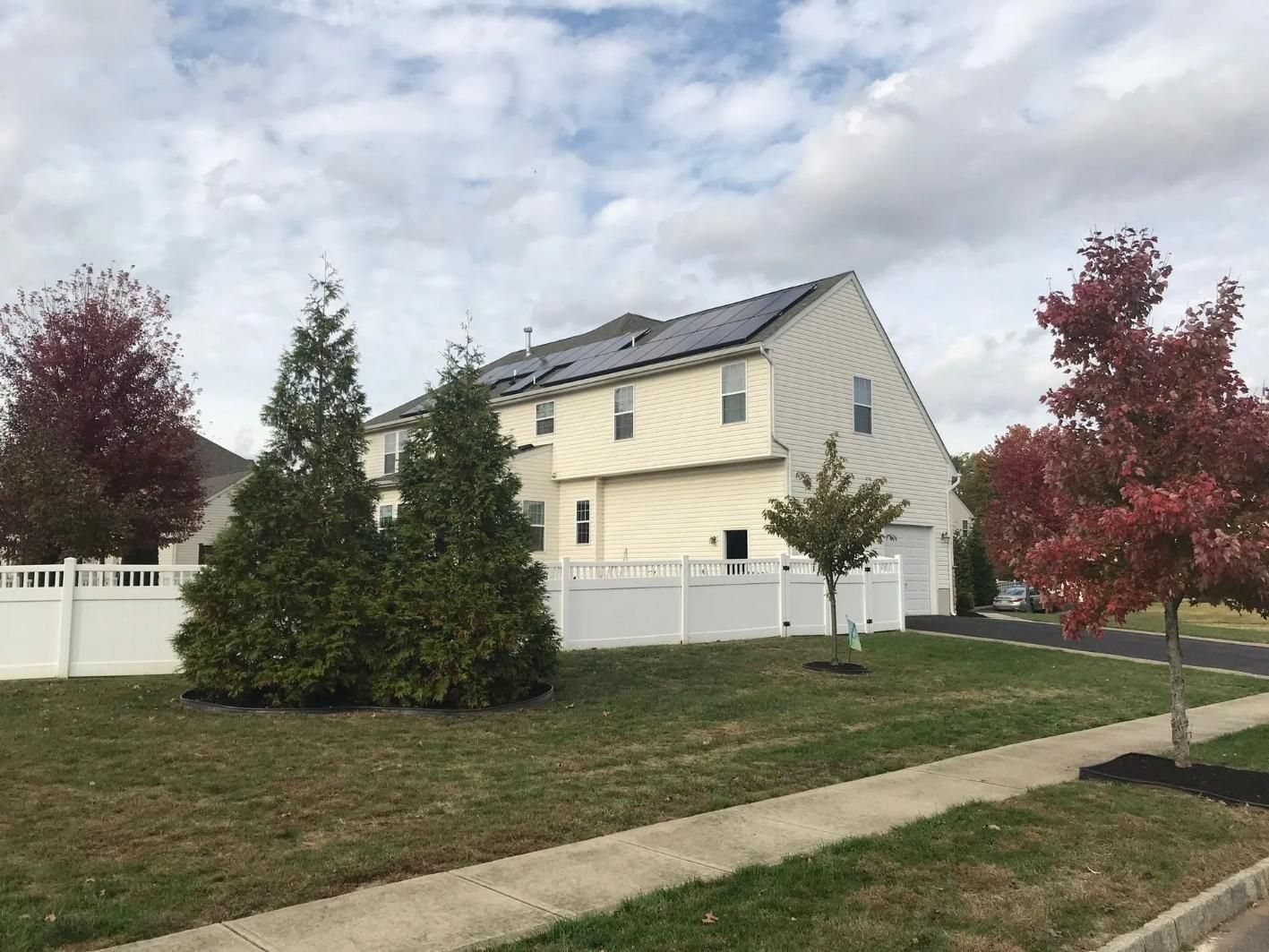 Two-story beige house with solar panels, white fence, trees with red leaves, cloudy sky.