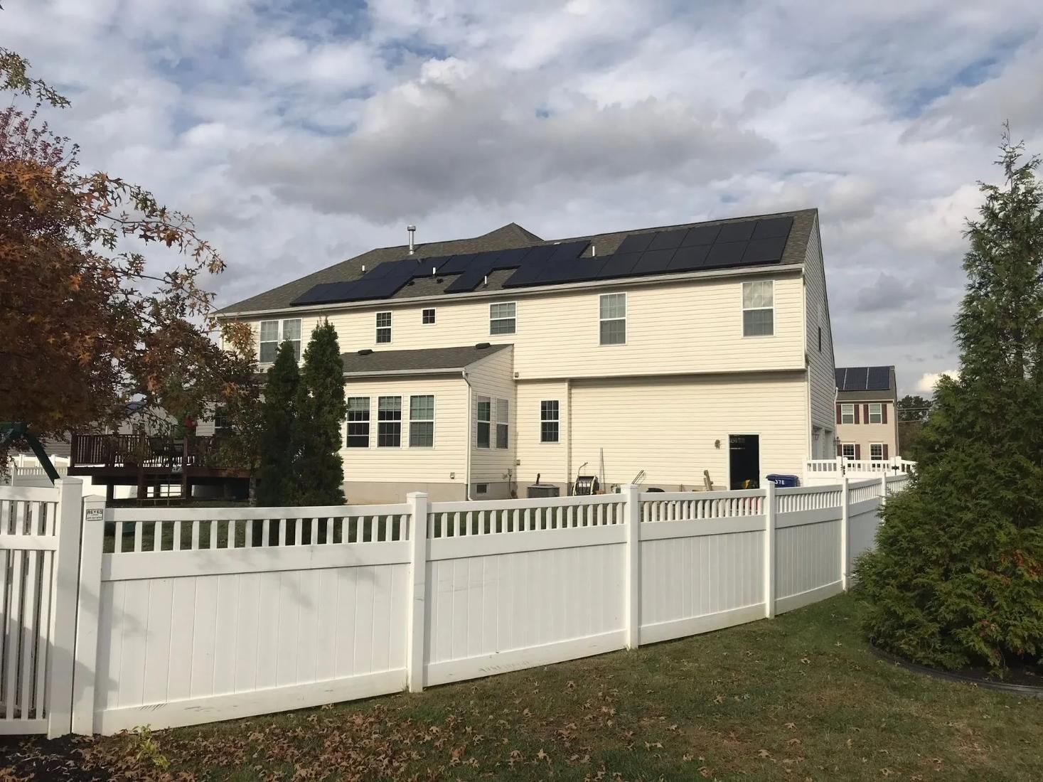 Back of a two-story beige house with solar panels on the roof and a white picket fence in the yard.