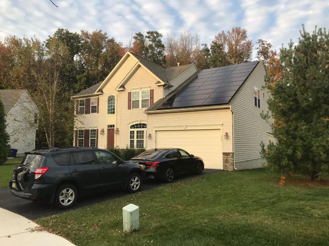 House with solar panels on roof, two cars parked in driveway, fall foliage.