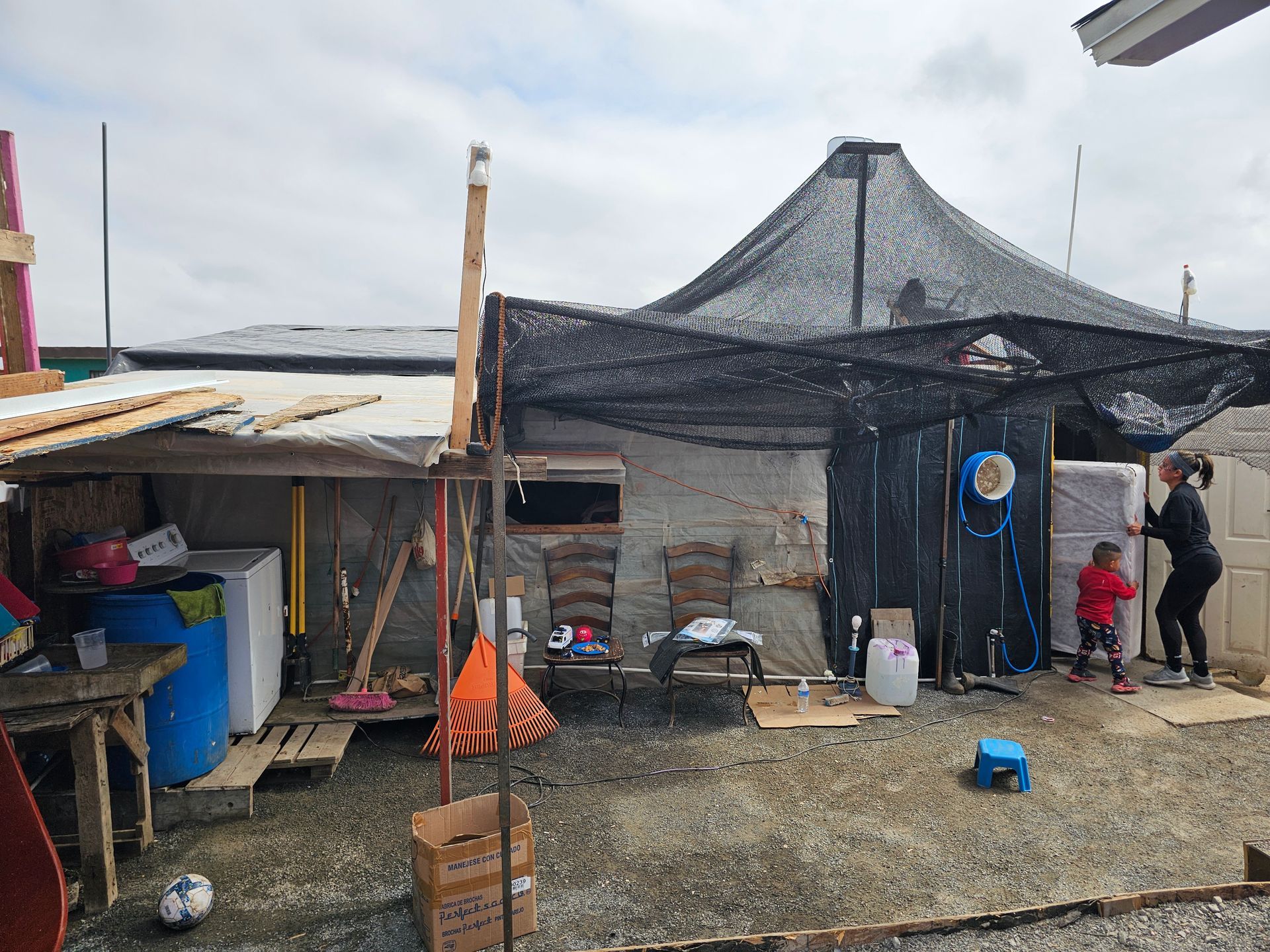 Small rustic outdoor shelter with tarps, netting, and tools; two people stand near a doorway on a gravel yard.