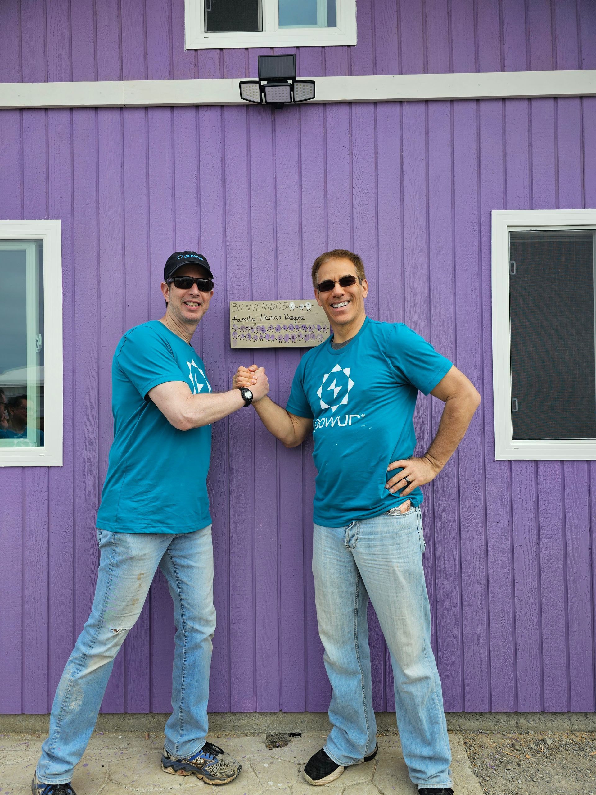 Two people in matching teal shirts stand in front of a purple building, posing with clasped hands.
