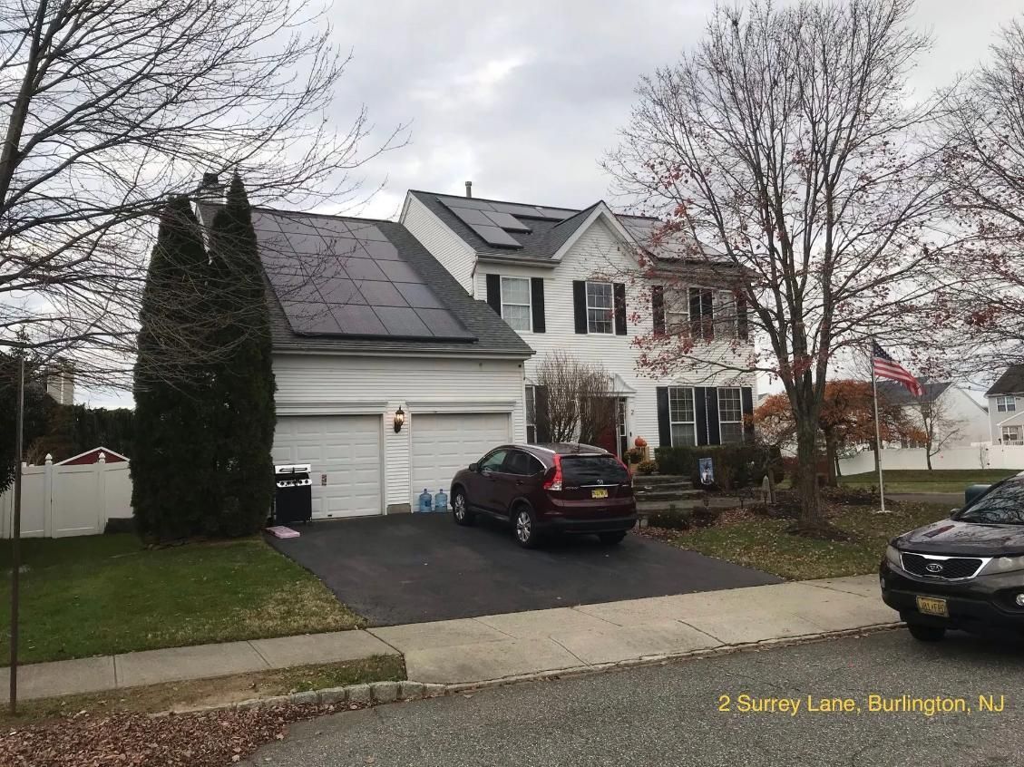 Two-story house with solar panels on roof, two-car garage, driveway with cars, and bare trees in suburban setting.