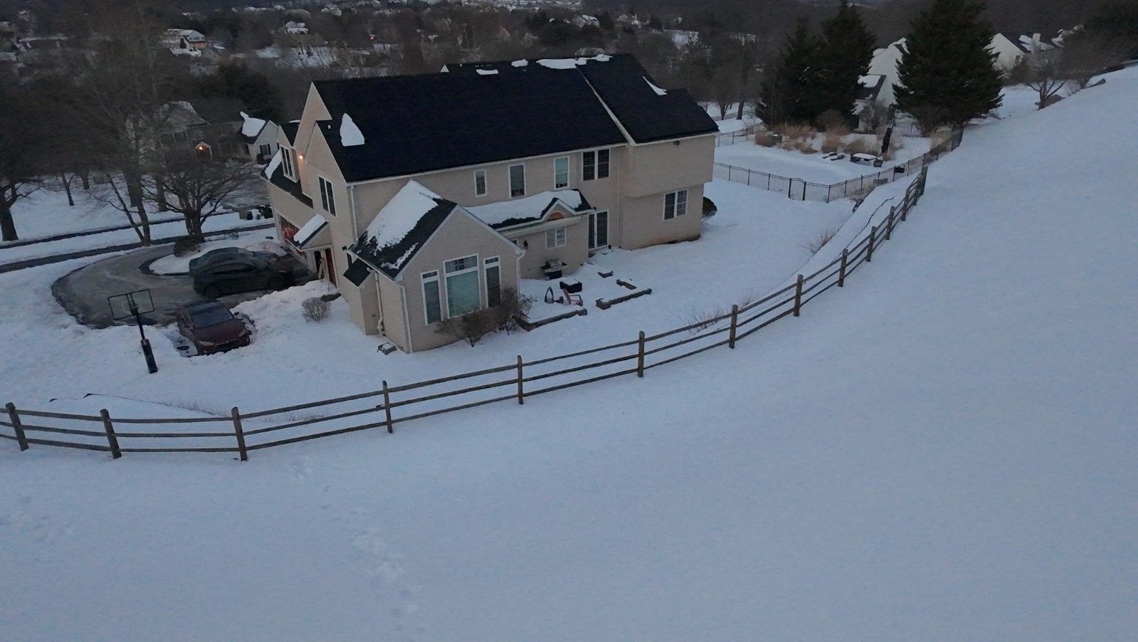 Snow-covered yard with a beige house, wooden fence, and trees in winter.