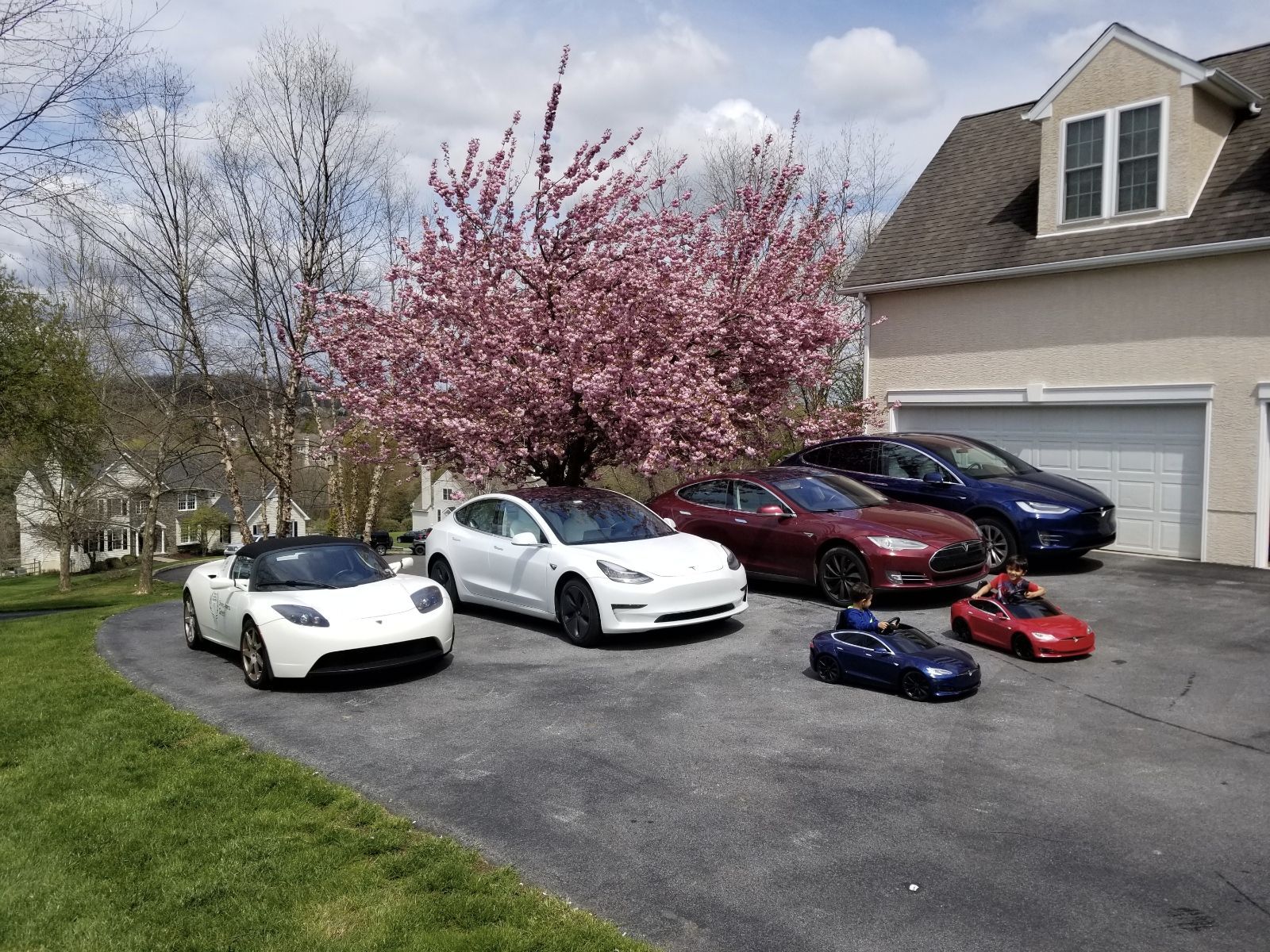 Several toy sports cars arranged in a driveway beside a house and blooming pink tree.
