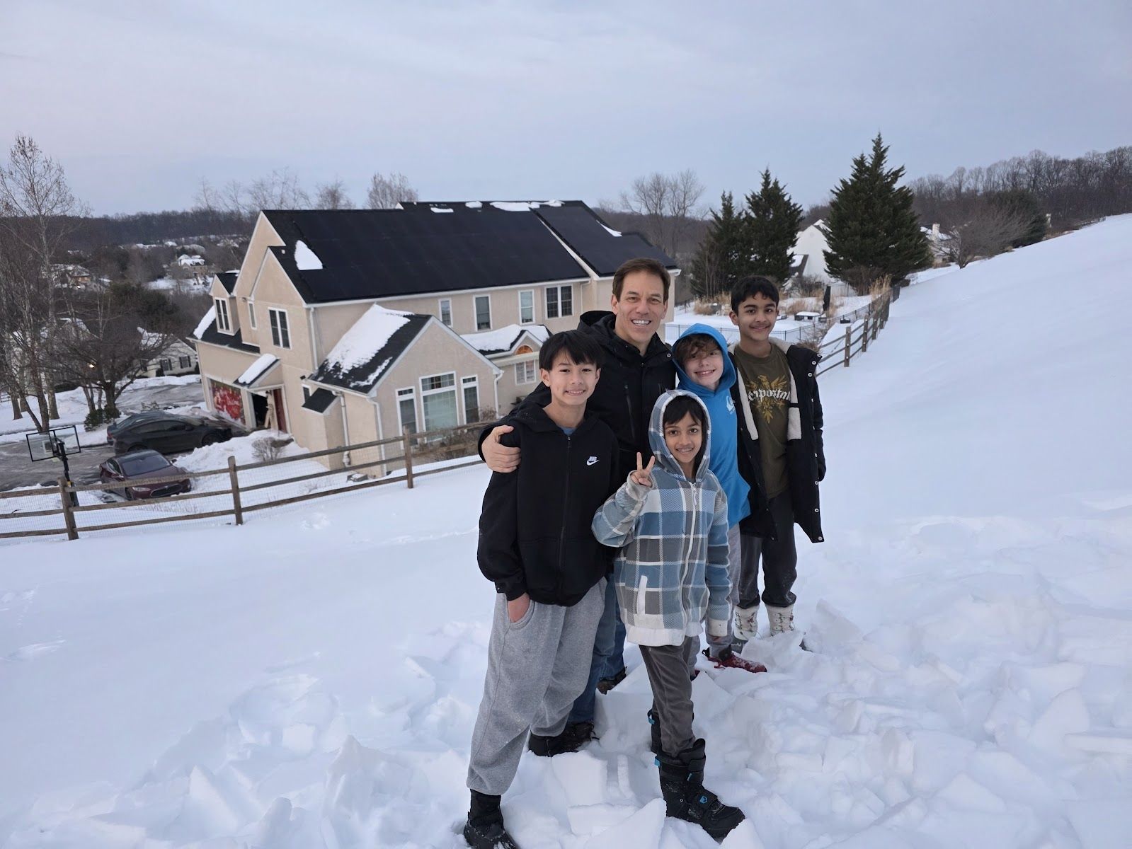Four people standing in snow near a house, with snowy hills and trees in the background.
