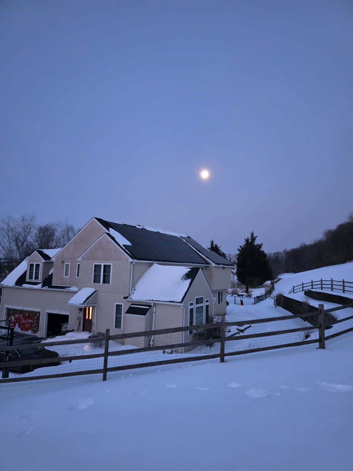 Snowy suburban houses at dusk under a pale moon, with a wooden fence in the foreground.