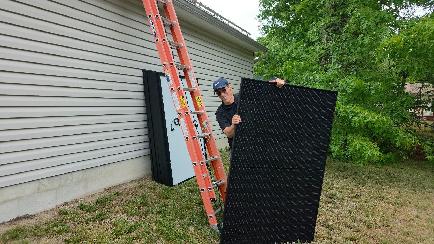Person stands by a ladder, carrying a large black panel beside a house.