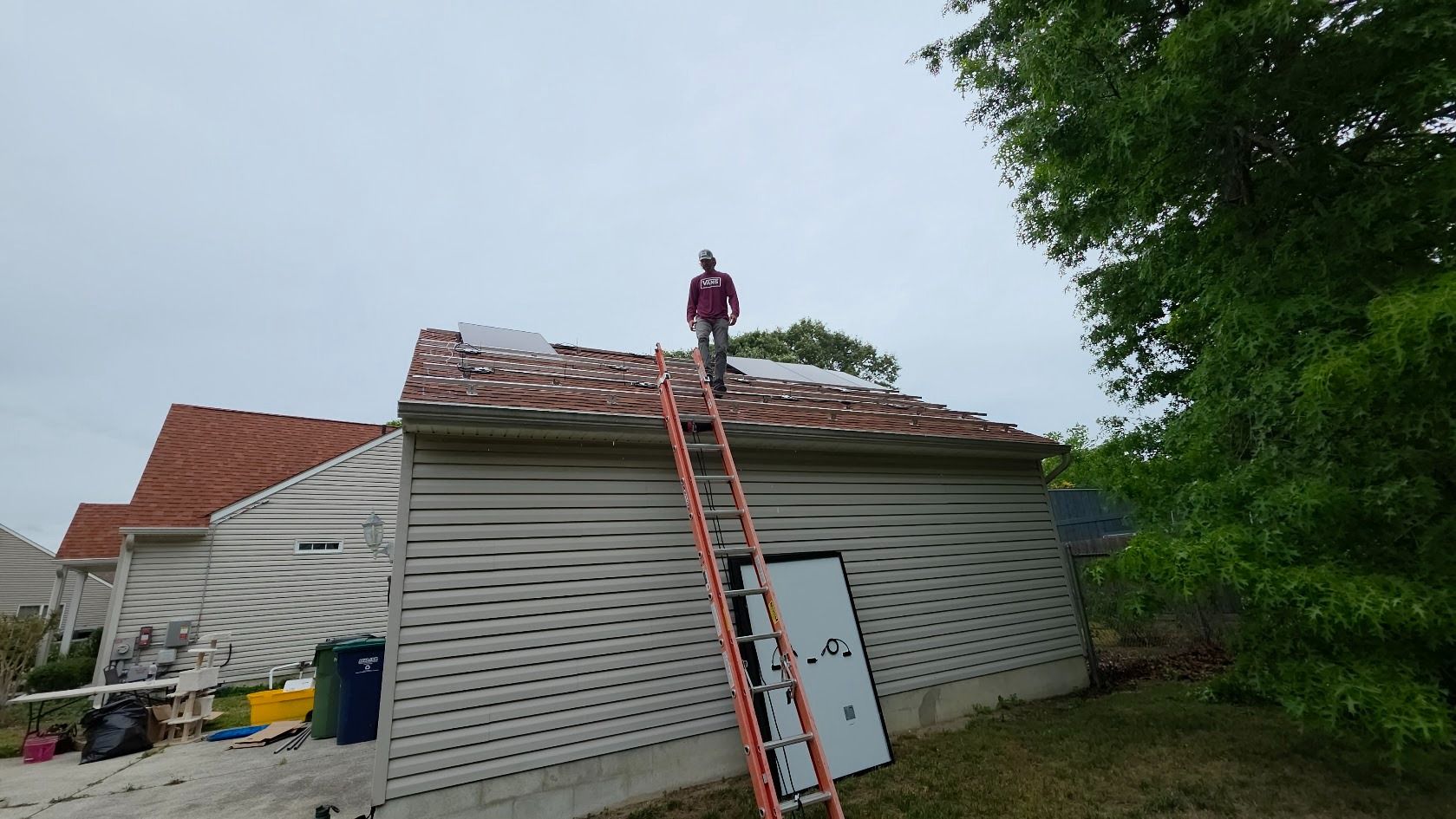 Worker on a roof with a ladder leaning to it, beside a house under renovation.