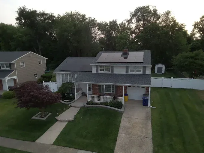 Two-story house with solar panels on roof, porch, driveway, and lush green lawn. Trees in the background.