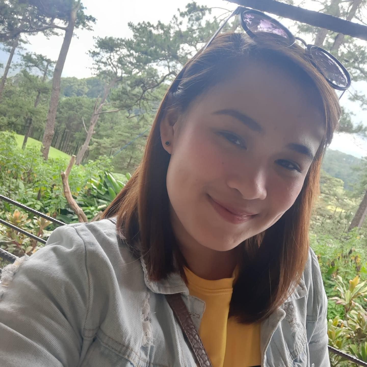 Woman with brown hair smiles outdoors, wearing sunglasses, yellow shirt, and denim jacket. Trees and greenery in background.