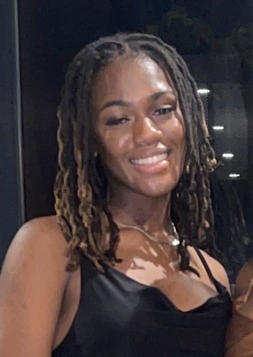 Woman with dreadlocks smiles, wearing a black satin top, outside.