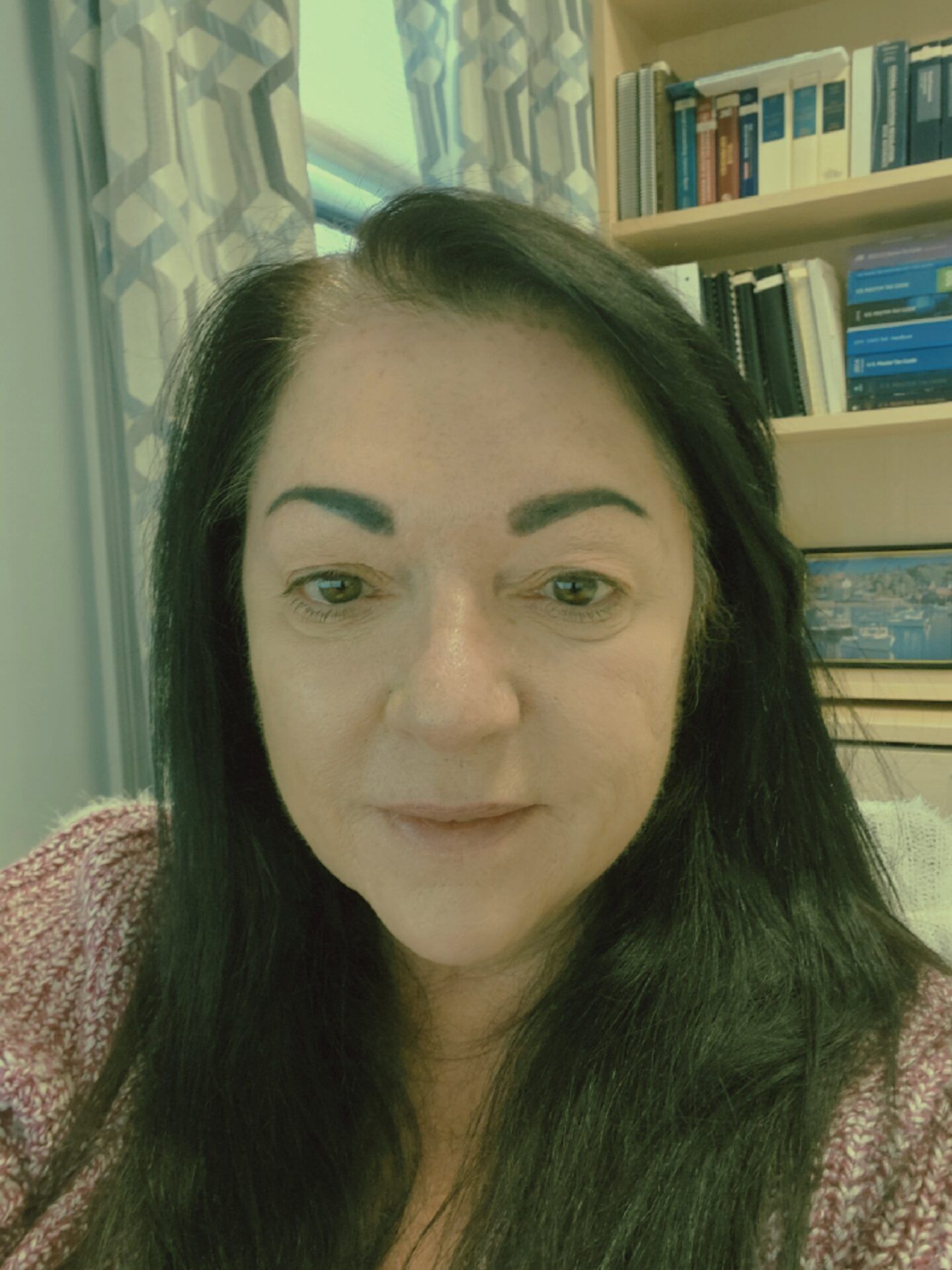 Woman with long dark hair, looking at the camera. In a room with bookshelves and a window.