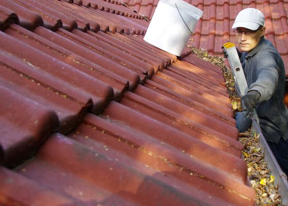 Worker cleaning gutters on a red tile roof with a vacuum tool.