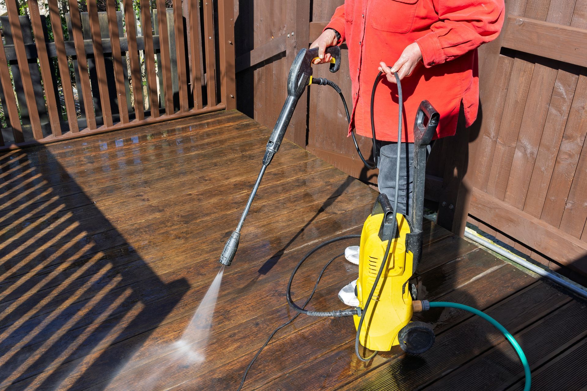 Person in red jacket pressure washing a wooden deck with a yellow machine.